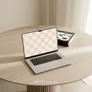 Op de afbeelding: Een zilveren laptop met een geruit patroon op het scherm staat op een lichtbruine houten tafel. De laptop is open en het toetsenbord is zichtbaar. Een zwart boek met witte tekst ligt rechts van de laptop op de tafel.