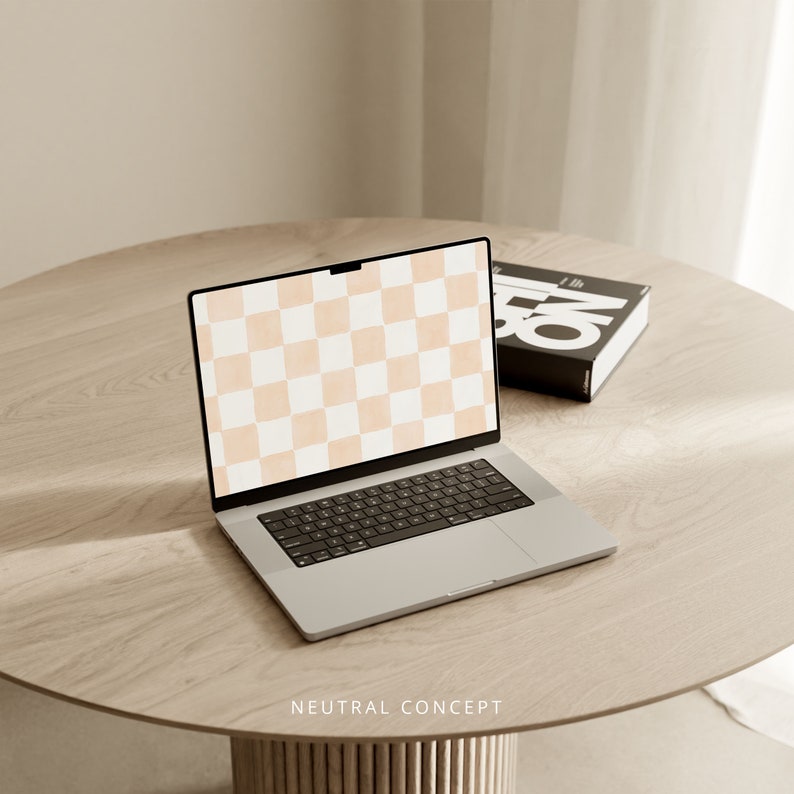Op de afbeelding: Een zilveren laptop met een geruit patroon op het scherm staat op een lichtbruine houten tafel. De laptop is open en het toetsenbord is zichtbaar. Een zwart boek met witte tekst ligt rechts van de laptop op de tafel.