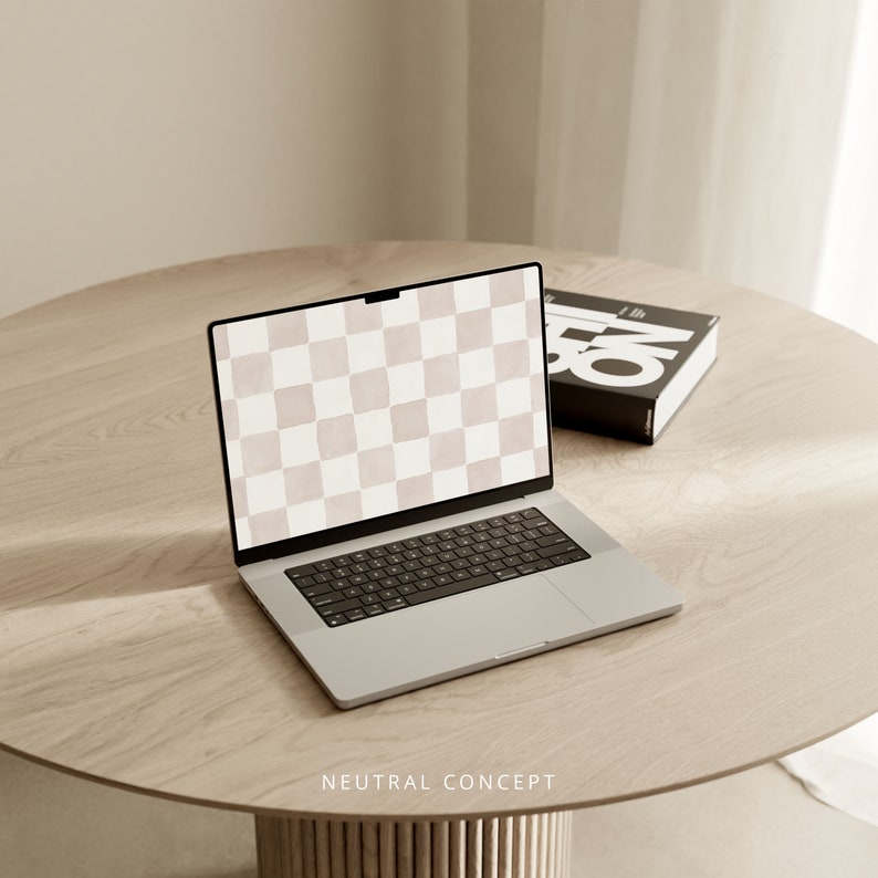 Op de afbeelding: Een zilveren laptop met een leeg scherm op een lichtbruine houten tafel. De laptop is open en het toetsenbord is zichtbaar. Een zwart boek met witte tekst ligt rechts van de laptop op de tafel.