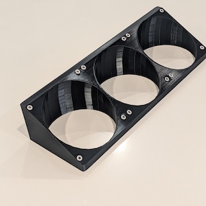 Puede incluir: Un objeto negro rectangular impreso en 3D con tres aberturas circulares. El objeto tiene forma trapezoidal y está asegurado con pequeños tornillos plateados. El diseño sugiere un soporte de montaje para artículos cilíndricos.