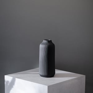 Könnte beinhalten: Eine schwarze Keramikvase mit vertikalen Rippen. Die Vase steht auf einer weißen Oberfläche.