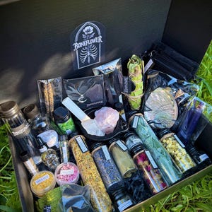 May include: Open black box filled with various spiritual items. Includes jars of herbs, crystals, a necklace, and a smudge stick. The box has a sticker with the text "Boneflower" and a floral and ribcage design.