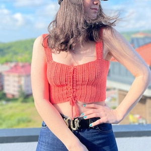 May include: A woman wearing a coral crochet top with a tie at the waist, blue jeans, and a black baseball cap. She is standing on a rooftop with a view of a city in the background.