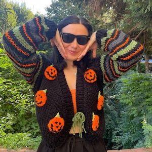May include: A black knitted cardigan with orange and green stripes and pumpkin appliques. The cardigan has a tie closure and is worn over a black tank top.