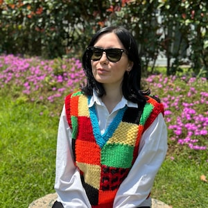 Può includere: Una donna indossa una camicia bianca e pantaloncini neri, con un gilet di maglia patchwork colorato. Indossa occhiali da sole neri ed è seduta su una pietra.