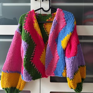 Pode incluir: Um cardigan de malha colorido com um design de patchwork em tons de rosa, verde, amarelo, azul e roxo. O cardigan tem um decote em V e mangas compridas.