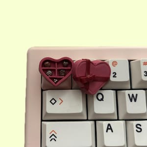 Puede incluir: Dos cajas de chocolate en forma de corazón, una con una cinta roja y la otra con una variedad de piezas de chocolate en su interior, se colocan en un teclado blanco con acentos rosados.