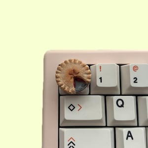 Puede incluir: Un tapón de tecla en miniatura con forma de pastel para un teclado mecánico. El tapón de tecla es marrón y tiene una corteza escamosa con un relleno rojo y negro. El tapón de tecla está en un teclado blanco con las teclas etiquetadas "!", "1", "@", "2", "< >", "Q" y "A".