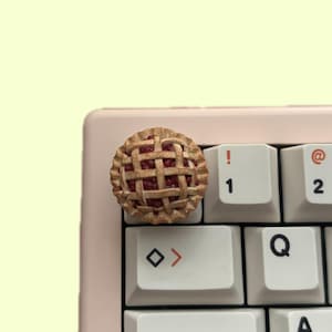 Może przedstawiać: Nakładka na klawisz klawiatury w kształcie wiśniowego ciasta. Nakładka jest wykonana z żywicy i ma kształt ciasta z kratką i czerwonym nadzieniem. Nakładka znajduje się na białym klawiszu klawiatury z symbolem "!" i cyfrą "1" nadrukowaną na nim.