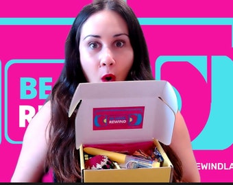 May include: A woman is holding open a box with the word "REWIND" on the top. The box is filled with various items, including a yellow hairbrush, a red and white striped scarf, and a small bottle of clear liquid. The background is a bright pink with a blue and yellow graphic.
