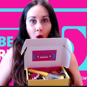 Puede incluir: Una mujer sostiene una caja abierta con la palabra "REWIND" en la parte superior. La caja está llena de varios artículos, incluyendo un cepillo para el cabello amarillo, una bufanda a rayas rojas y blancas y una pequeña botella de líquido transparente. El fondo es de color rosa brillante con un gráfico azul y amarillo.