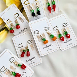 Peut inclure: Une collection de boucles d'oreilles faites à la main représentant divers fruits et légumes. Les boucles d'oreilles sont présentées sur des cartes blanches avec des crochets dorés et le texte "skaybystebby cute jewelry". Les boucles d'oreilles comprennent des cerises rouges, des olives noires et des citrouilles oranges.