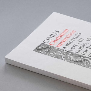 May include: A white book with black and gray text and a decorative design on the cover. The text reads "CIMUS Christum surrexisse a mortuis vere: tu nobis, vic-tor Rex, miserere. Amen. Alleluia."