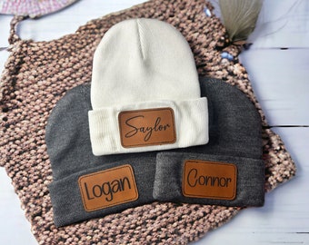 May include: Three knit beanies with personalized leather patches. The beanies are gray and white. The patches are brown with the names "Saylon", "Logan", and "Connor" written in black.