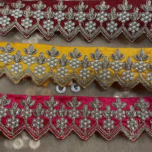 Puede incluir: Tres filas de ribete decorativo con borde festoneado. El ribete está bordado con un diseño floral y adornado con pequeñas cuentas blancas. La fila superior es burdeos, la fila del medio es amarilla y la fila inferior es rosa.