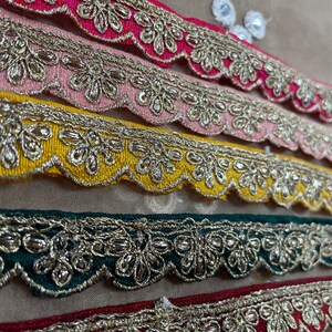 Op de afbeelding: Close-up van decoratieve sierranden in verschillende kleuren, waaronder roze, geel, groen en rood. Elke rand heeft een geschulpte rand en ingewikkelde gouden metalen bloemenborduursels. De randen zijn in parallelle rijen gerangschikt en tonen hun gedetailleerde ontwerp.