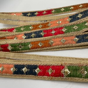 May include: Multicoloured trim with gold sequins and a beige backing. The trim features a repeating pattern of red, orange, green, and blue squares.