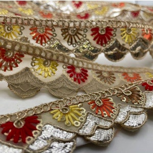 May include: Close-up of decorative trim with a scalloped edge. The trim features embroidered floral designs in red, yellow, and orange, with gold and silver sequins. The trim is beige and has a delicate, lacy appearance.