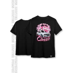 以下が含まれることがあります： ピンクのグラフィックがプリントされた黒いTシャツ。白い車がピンクのガレージの前にあり、ヤシの木と「私は年をとっていない、私はクラシックだ」というテキストが書かれている。