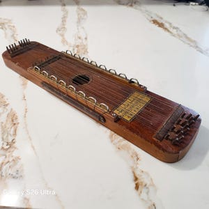 May include: A rectangular wooden zither stringed instrument featuring a dark brown finish, multiple metal tuning pins at both ends, a central circular sound hole, and a gold-colored metal chord chart plate with printed musical notation and numbers.