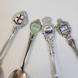 May include: Four decorative souvenir spoons with detailed handles and colorful enamel crests. The spoons feature designs from London, Niagara Falls, Puerto Rico, and Brussels. The spoons are made of silver-colored metal and are displayed on a white surface.