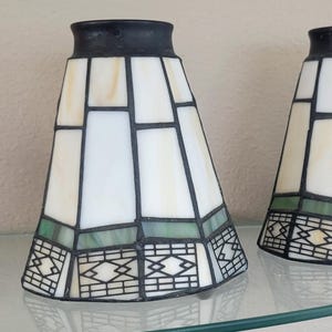 May include: Two conical lamp shades constructed from individual panels of white and cream-colored glass joined by dark metal soldering. Each shade features a horizontal band of green glass near the base and a geometric black metal pattern.