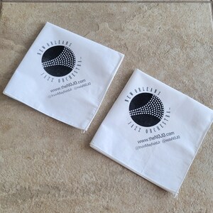 May include: Two white fabric squares with black text and a logo. The logo is a black circle with a stylized trumpet inside. The text reads "New Orleans Jazz Orchestra" and includes the website and social media handles.