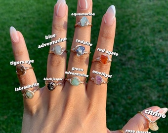 May include: A collection of wire-wrapped rings featuring raw gemstones including aquamarine, sodalite, red agate, amethyst, green aventurine, black jasper, tigers eye, labradorite, citrine, and rose quartz. The image includes the text ALL SIZES AVAILABLE!!!