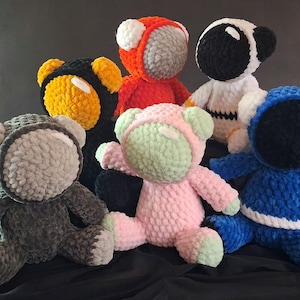 May include: A collection of handmade crochet astronaut bears in various colors, including black, white, red, blue, pink, and gray. Each bear has a unique helmet design and is made with soft, textured yarn. The bears are arranged on a dark background.