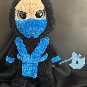 Könnte beinhalten: Handgefertigte Plüschpuppe eines Ninja-Charakters mit schwarzem Kapuzenoutfit, blauer Maske und passenden Akzenten. Die Puppe hat große, ausdrucksstarke Augen und ein hellbraunes Gesicht. Sie hält eine durchscheinende blaue Axt.