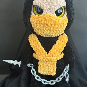 May include: A handmade plush doll of a character in a black hooded outfit with a yellow mask and accents. The doll has large yellow eyes and a silver chain and dagger. The doll is approximately 12 inches tall.