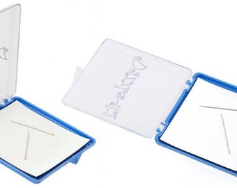 May include: A clear plastic case with a blue interior and a white paper insert. The case is open and two sewing needles are visible on the paper. The case has the text "Dritz-Craft" printed on the outside.