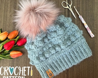 May include: A light blue crochet beanie with a fluffy pink pom pom. The beanie has a textured stitch pattern and a brown leather tag with a logo.