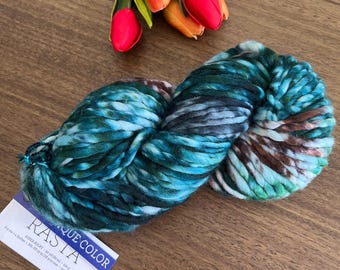 May include: A skein of super bulky Rasta yarn in a variegated blend of teal, dark green, black, and brown, accompanied by a white product label with black text and three artificial tulips in shades of red, orange, and yellow.