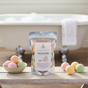 May include: A clear plastic bag filled with colorful mini bath bombs, labeled "Rainbow Sherbet." A small bowl holds additional bath bombs in pink, orange, yellow, and green. The scene is set on a wooden surface with a blurred background of a bathtub.