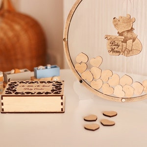 以下が含まれることがあります： ハート型のカットアウトデザインと「A Little Bit Sweet Baby Shower」という刻印が施された木製の箱。背景には、くまのプーさんのキャラクターと「Baby Mason」という文字が刻まれたハート型の木製ゲストブックがあります。