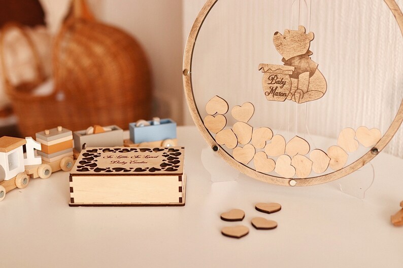 以下が含まれることがあります： ハート型のカットアウトデザインと「A Little Bit Sweet Baby Shower」という刻印が施された木製の箱。背景には、くまのプーさんのキャラクターと「Baby Mason」という文字が刻まれたハート型の木製ゲストブックがあります。
