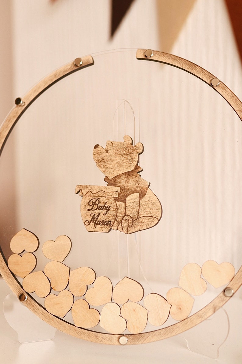 以下が含まれることがあります： 透明なアクリル製のフレームが付いた、木製のかわいいプーさんのベビーシャワーのゲストブックです。フレームには、ゲストがサインできる小さな木製ハートが詰められています。プーさんのキャラクターは、蜂蜜の壺を持っており、「Baby Mason」という文字が刻まれています。