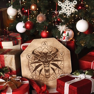 K&ouml;nnte beinhalten: Ein h&ouml;lzerner Weihnachtsbaumschmuck mit einem Wolfs Kopf und einem Symbol eines Wolfs Kopfes in einem Kreis. Der Schmuck ist umgeben von verpackten Geschenken und einem Weihnachtsbaum mit wei&szlig;en und roten Kugeln.