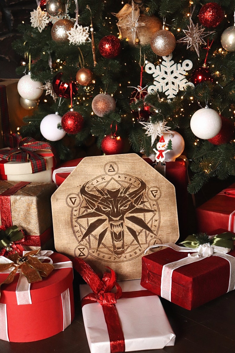 K&ouml;nnte beinhalten: Ein h&ouml;lzerner Weihnachtsbaumschmuck mit einem Wolfs Kopf und einem Symbol eines Wolfs Kopfes in einem Kreis. Der Schmuck ist umgeben von verpackten Geschenken und einem Weihnachtsbaum mit wei&szlig;en und roten Kugeln.