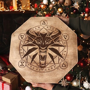 K&ouml;nnte beinhalten: Ein achteckiges Holzschild mit einem Wolfskopf und einem Symbol eines Wolfskopfes in einem Kreis mit Runen drumherum. Das Schild wird von einer Person in der Hand vor einem Weihnachtsbaum gehalten.