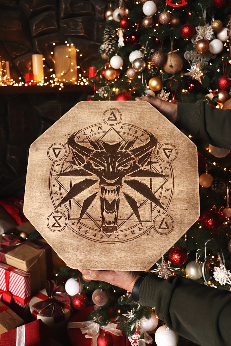 K&ouml;nnte beinhalten: Ein achteckiges Holzschild mit einem Wolfskopf und einem Symbol eines Wolfskopfes in einem Kreis mit Runen drumherum. Das Schild wird von einer Person in der Hand vor einem Weihnachtsbaum gehalten.