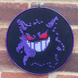 May include: A round, black fabric embroidery hoop featuring a pixelated purple and white Gengar design with red eyes. The hoop has a purple border and a metal clasp.