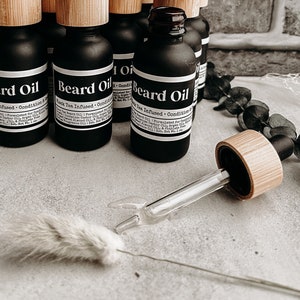 May include: A close-up of multiple small black glass bottles with bamboo lids and droppers. The bottles are labeled "Beard Oil" and "Black Tea Infused - Condition & Soften".