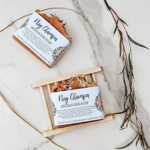 Puede incluir: Dos barras de jabón artesanal Nag Champa, cada una con un diseño floral y el texto "Nag Champa Artisan Bar Soap" y una lista de ingredientes. Las barras de jabón están en un jabonera de madera.