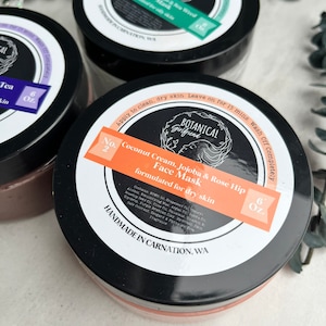 May include: Three jars of face mask products with black lids and colorful labels. The labels read "Botanical Girlfriend Face Mask" with different ingredients and sizes. The jars are labeled "Coconut Cream, Jojoba & Rose Hip", "Charcoal, Minerals & Sea Weed", and "Green Tea".