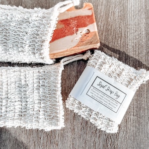 May include: Two white sisal soap bags with a bar of soap in one of the bags. The soap is a light brown, orange, and white color. The bag has a label that reads "Sisal Soap Bag" and instructions for use.