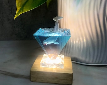 May include: A white whale figurine is encased in a clear triangular resin block with a blue liquid. The block sits on a wooden base with a small light underneath.