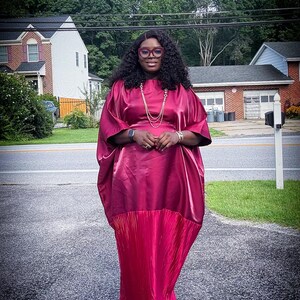 May include: A burgundy-colored dress with a flowing, pleated skirt and a loose-fitting top. The dress has a satin-like sheen and is worn with glasses and jewelry. The background includes a residential street and houses.