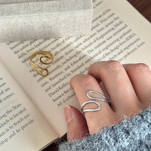 May include: A gold-toned open-style ring and a silver-toned open-style ring, both featuring a fluid, wave-like design. The gold ring is positioned near a rectangular beige fabric ring display stand, while the silver ring is worn on a finger.